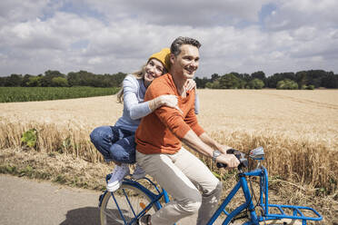 Lächelnde Frau umarmt Mann auf Fahrrad vor einem Feld an einem sonnigen Tag - UUF29683