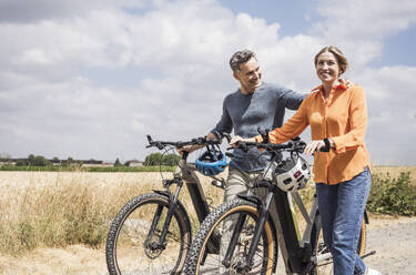 Happy couple wheeling bicycles near field on sunny day - UUF29713