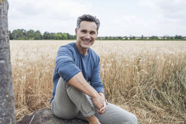 Smiling man sitting on rock near field - UUF29720