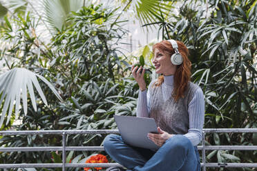 Glückliche Geschäftsfrau mit drahtlosen Kopfhörern, die mit einem Laptop sitzt und über ein Smartphone spricht - PNAF05893