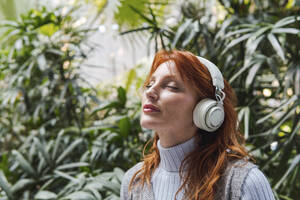 Frau hört Musik mit drahtlosen Kopfhörern vor Pflanzen - PNAF05896