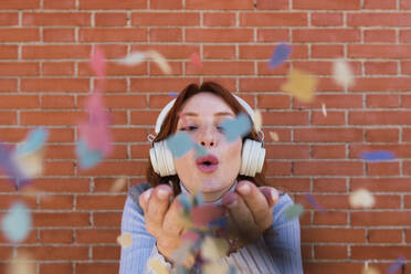 Woman wearing wireless headphones blowing confetti - PNAF05911