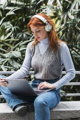 Geschäftsfrau mit drahtlosen Kopfhörern bei der Arbeit am Laptop - PNAF05931