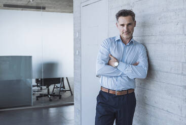 Smiling man with arms crossed leaning on wall in office - UUF29822