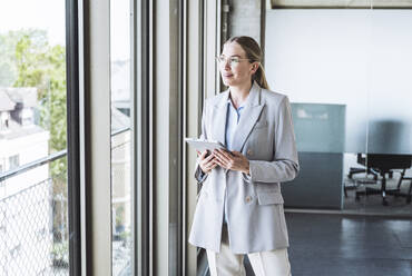 Thoughtful businesswoman standing with tablet PC by window in office - UUF29828