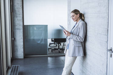 Lächelnde Geschäftsfrau mit Tablet-PC und an die Wand gelehnt im Büro - UUF29834