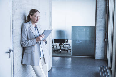 Smiling businesswoman using tablet PC leaning on wall in corridor - UUF29835