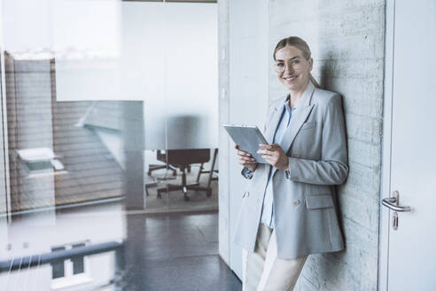 Happy businesswoman leaning on wall with tablet PC - UUF29836