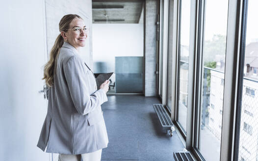 Happy businesswoman with tablet PC standing in office corridor - UUF29838