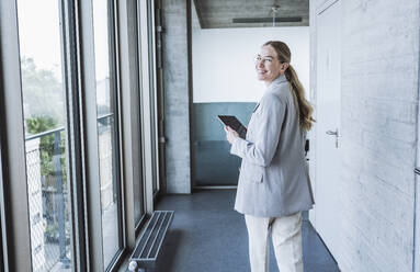 Happy businesswoman walking with tablet PC in corridor - UUF29839