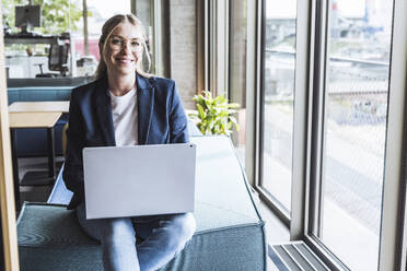 Happy businesswoman sitting with laptop in office - UUF29842