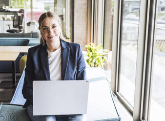 Smiling blond businesswoman sitting with laptop in office - UUF29843