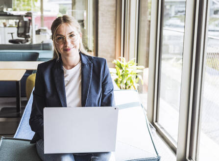 Lächelnde blonde Geschäftsfrau sitzt mit Laptop im Büro - UUF29843