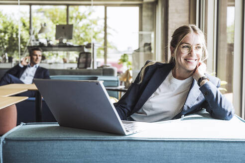 Happy businesswoman sitting with laptop on sofa in office - UUF29845