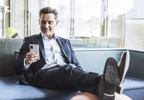 Lächelnder reifer Geschäftsmann mit Smartphone auf dem Sofa im Büro - UUF29848