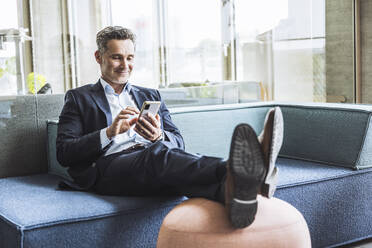 Lächelnder Geschäftsmann mit Smartphone auf dem Sofa im Büro - UUF29849