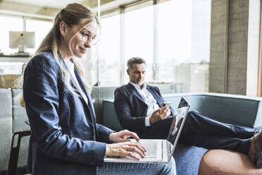 Lächelnde Geschäftsfrau, die an einem Laptop arbeitet, mit einem Geschäftsmann, der ein Smartphone im Hintergrund benutzt - UUF29853