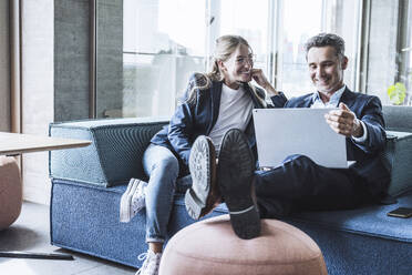 Smiling businessman and businesswoman having discussion on sofa and using laptop in office - UUF29858