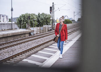 Woman wearing wireless headphones walking on railroad station platform - UUF29867