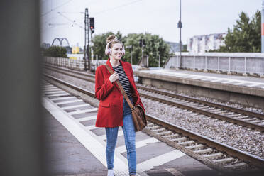 Lächelnde Frau mit drahtlosen Kopfhörern auf dem Bahnsteig eines Bahnhofs - UUF29868