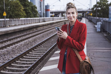 Happy woman standing with mobile phone at railroad station - UUF29877