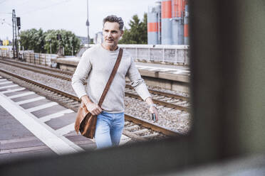 Smiling man with shoulder bag walking at railroad station - UUF29878