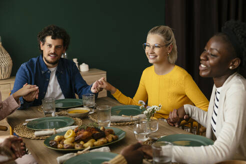 Happy friends holding hands and praying at dining table - EBSF03719