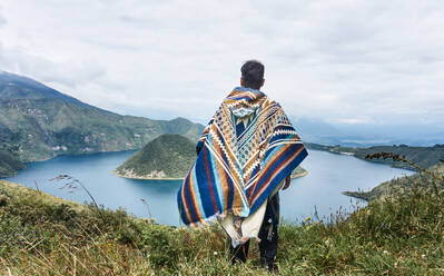 Back view of anonymous male traveler in wool hooded poncho looking away while standing on grassy hill and admiring scenery of Cuicocha lagoon Ecuador - ADSF46036