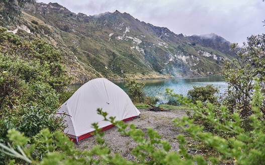 Scenic view of camping tent placed on rocky hill near Quilotoa lagoon Ecuador and surrounded by green trees mountains in daylight under cloudy sky - ADSF46037