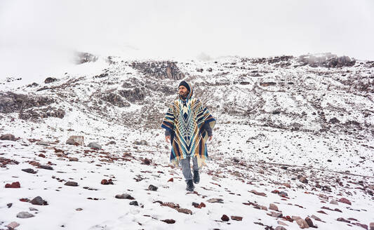 Full body of young bearded male traveler wearing hooded poncho looking away while walking on Chimborazo Volcano Ecuador against overcast sky during winter vacation - ADSF46041