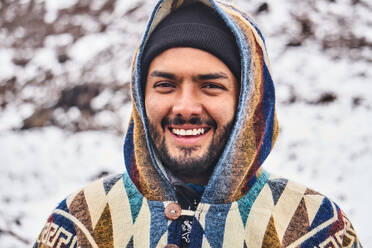 Portrait of positive bearded young male in poncho hood smiling and looking at camera while standing on blurred Chimborazo Volcano of Ecuador in winter - ADSF46042