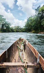 Old wooden boat with rope tied from bow to wooden plank floating in rippling blue river water of Ecuador near green trees against blue cloudy blue sky - ADSF46047