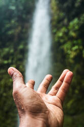 Crop hand of person in open palm gesture against waterfall in green tropical forest on sunny day in Ecuador - ADSF46050