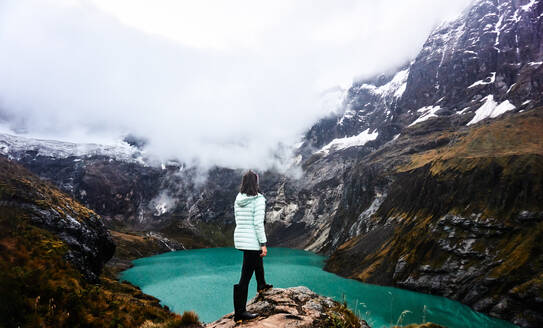 Back view of anonymous female traveler in cozy clothes standing on rocky cliff and admiring scenery view of cloudy mountains and lake in valley with volcano Altar in Ecuador - ADSF46054