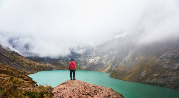 Back view of unrecognizable male traveler in warm clothes standing on edge of cliff and admiring scenic river and cloudy mountains of volcano Altar in Ecuador - ADSF46055