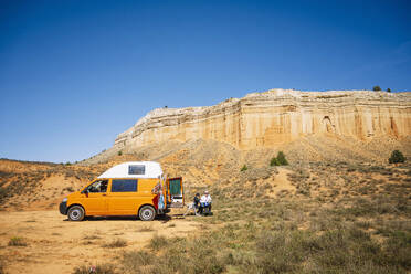 Modern camper van parked with people sitting on grassy terrain near hilly ridge against cloudless blue sky on sunny day at canyon Rojo Teruel Spain - ADSF46059