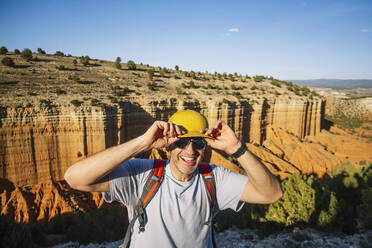 Happy mature male tourist in sunglasses and casual clothes with backpack touching cap while standing on edge of canyon and looking at camera - ADSF46065