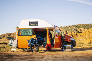 Älterer nachdenklicher Mann sitzt auf einem klappbaren Sessel und trinkt Kaffee neben einem modernen Wohnmobil, das auf einer sandigen Ebene einer Bergschlucht an einem sonnigen Tag geparkt ist - ADSF46067