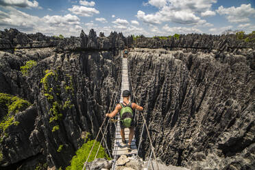 Back view of unrecognizable bare shouldered male hiker with backpack walking on wooden plank ropeway bridge of national park Madagascar Africa - ADSF46074