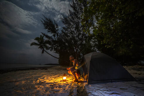 Side view of man sitting near tent with burning lamp and looking at sea while resting on sandy beach of Madagascar at night time - ADSF46081