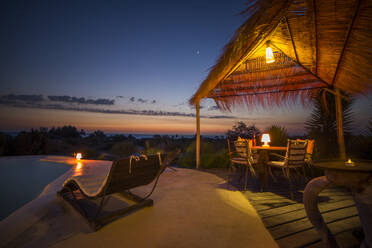 Illuminated poolside with bungalow pergola and table with chairs against ocean in evening time, Madagascar - ADSF46087