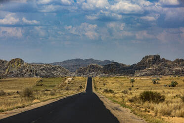 Empty asphalt road going through dried up bushes and small trees in Madagascar countryside with rocky mountains against blue cloudy sky in daylight - ADSF46095