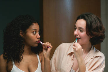 Calm young multiracial lesbian females with dark hair standing and looking at each other while brushing teeth together near mirror in illuminated room - ADSF46114