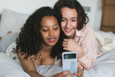 Junge afroamerikanische Frau mit lächelnder, vielseitiger Freundin, die ihre lockigen Haare berührt und gemeinsam auf den Bildschirm ihres Mobiltelefons schaut, während sie auf einem weißen Bett zu Hause liegt und das Wochenende genießt - ADSF46120
