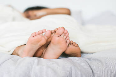 Crop anonymous bare feet of loving lesbian couple lying under white blanket in cozy bed at home against blurred background - ADSF46124