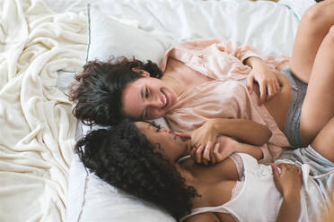 High angle of cheerful multiracial lesbian couple lying on bed and holding hands while laughing and enjoying romantic moments in daytime - ADSF46125