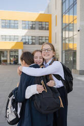From below of smiling young adolescent schoolgirls with backpacks looking at camera while standing and hugging unrecognizable classmate near building - ADSF46128