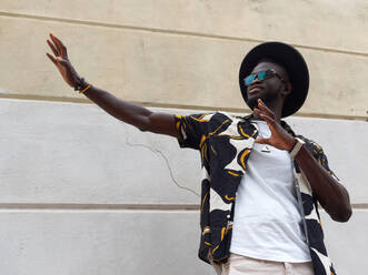 From below of delighted young African American guy in trendy outfit glasses and hat smiling and gesturing while standing against white wall on street - ADSF46137