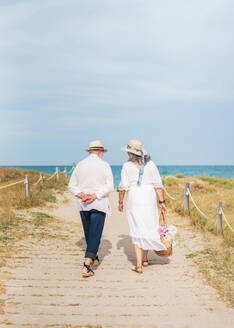 Rückenansicht eines älteren Mannes und einer anonymen Frau in Strandkleidung und Hüten, die unter hellem Sonnenlicht auf dem Weg zum Meer gehen - ADSF46142