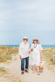Glücklicher älterer Mann und Frau in Strandkleidung und Hüte zu Fuß auf dem Weg den Strand unter hellem Sonnenlicht - ADSF46143